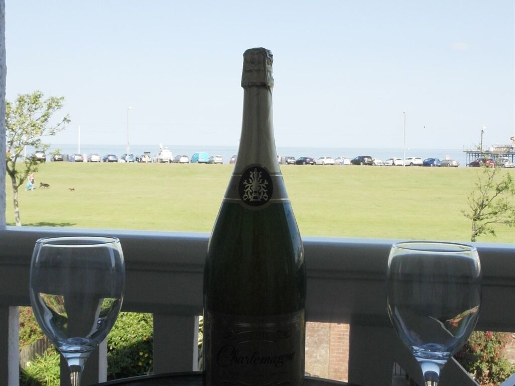 A bottle of champagne on a balcony with two empty glasses, overlooking a grassy area with parked cars and a clear sky.