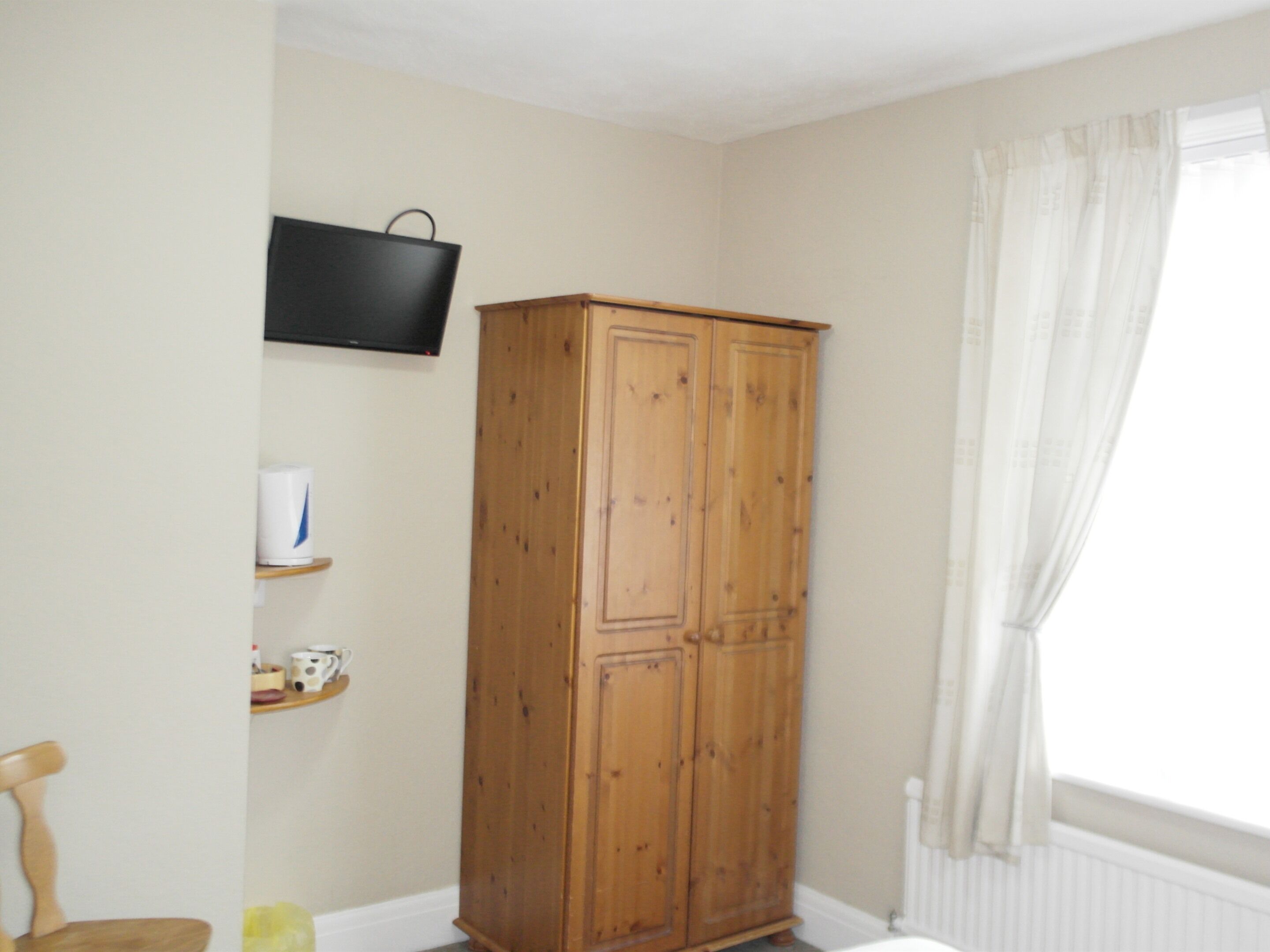 Une pièce avec une armoire en bois, un téléviseur fixé au mur et une fenêtre avec des rideaux.