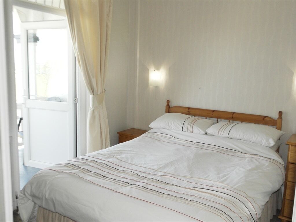 A bedroom with a wooden bed frame, white bedding, a nightstand, and a wall-mounted light.