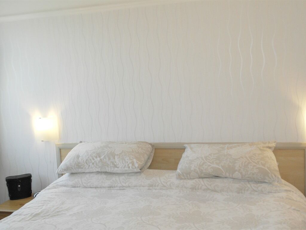 Un lit avec deux oreillers et une tête de lit, une table de chevet avec une lampe, et un mur orné d’un motif texturé.
