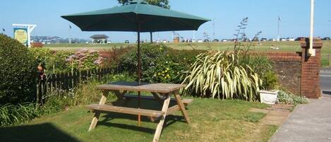 A picnic table with a green umbrella, surrounded by a grassy area with flowers and a brick wall.