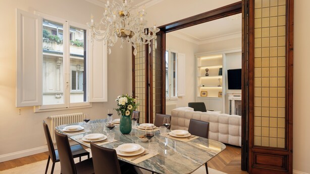 Un coin repas avec une table en marbre, des chaises et un lustre. On aperçoit un canapé et un téléviseur à l’arrière-plan.