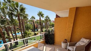 A balcony with a table and chairs, a small table with two cups, a potted plant, and a view of a pool and palm trees.