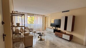 A spacious living room with a marble floor, a glass coffee table, a sofa, and a TV mounted on the wall.