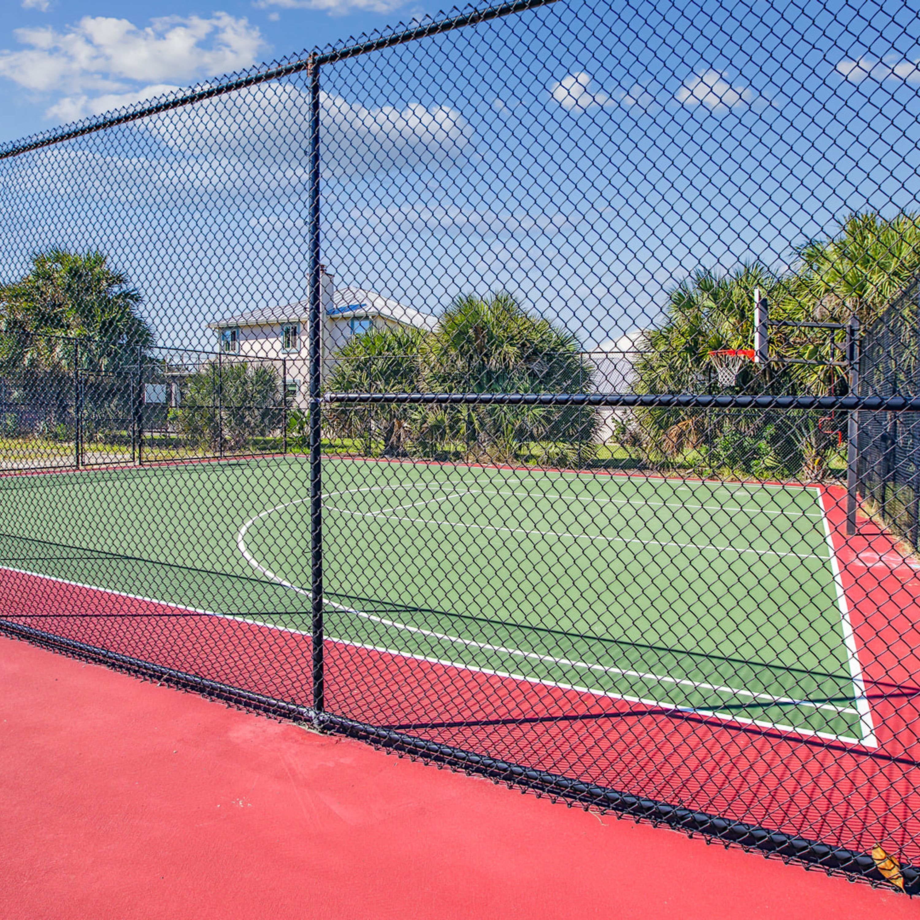 A tennis court enclosed by a chain-link fence with a red surface and a green boundary.