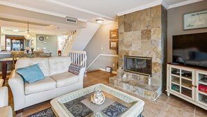 A spacious living room with a stone fireplace, a television, and a dining area with a kitchen in the background. - Oceanfront 3BR Luxe New Rental (St. Augustine)