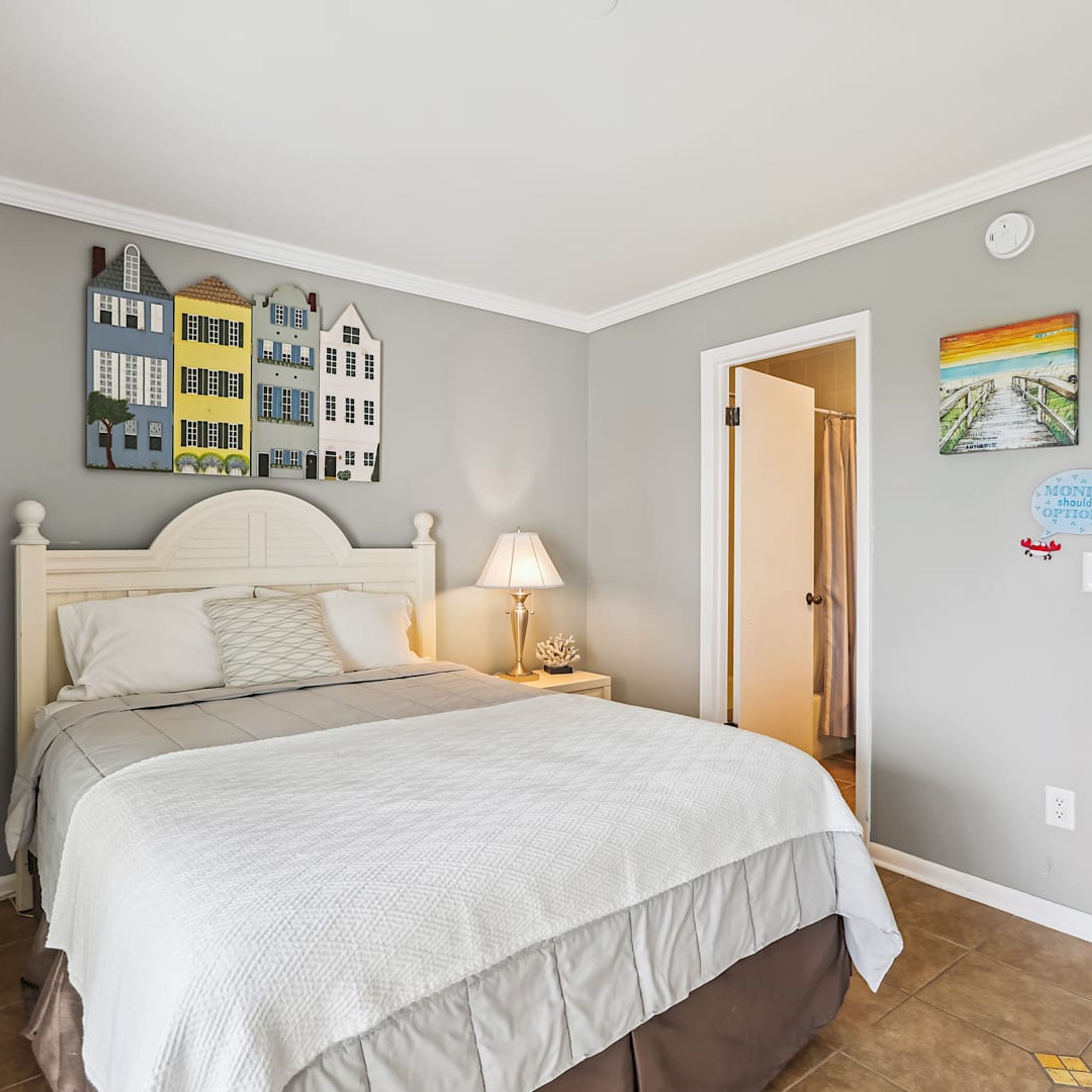 A neatly made bed with a white and gray duvet, a bedside lamp, and a wall with a series of colorful house cutouts.