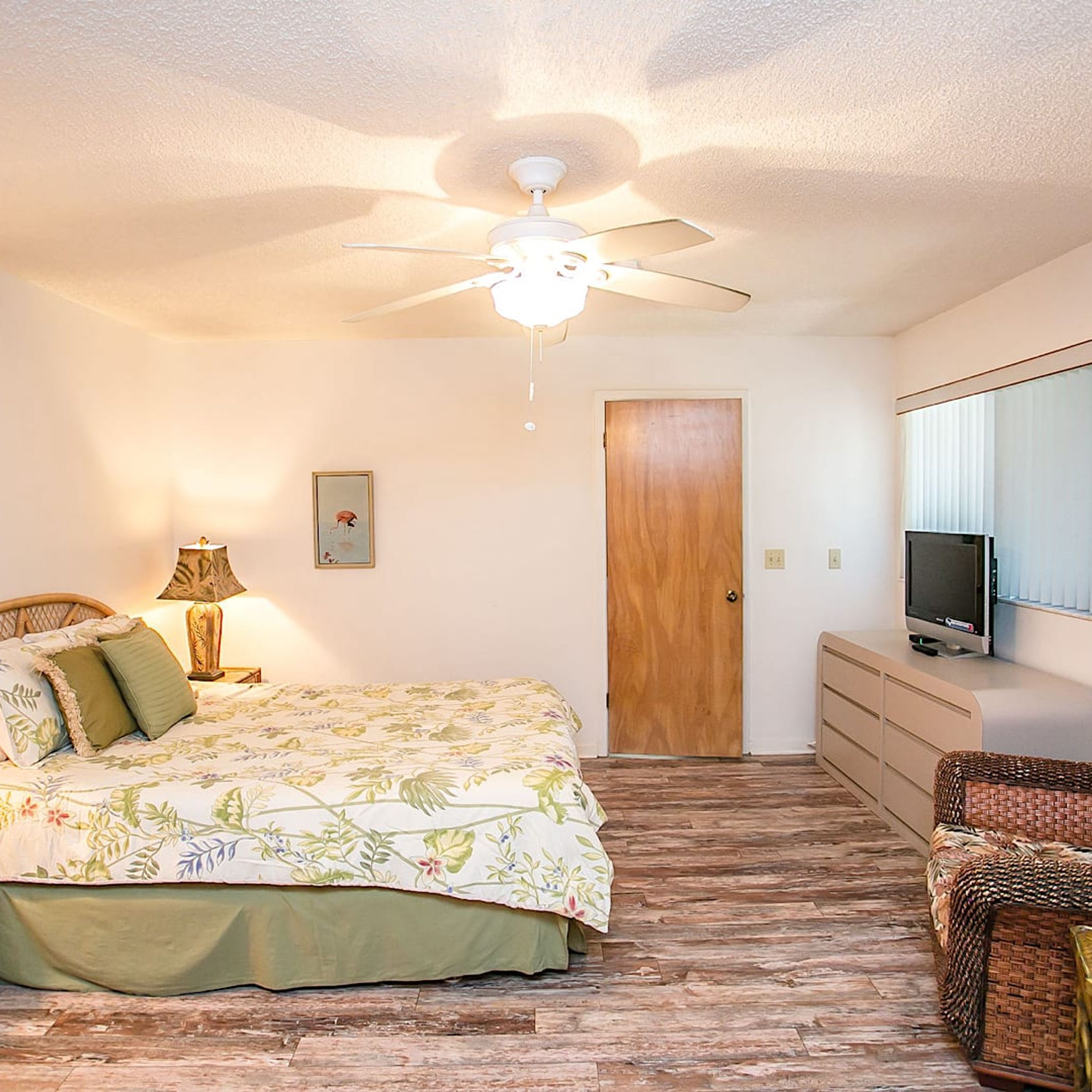 A bedroom with a bed, a ceiling fan, a wooden door, a television, and a wicker chair.