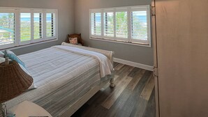 Ein Schlafzimmer mit einem Bett, einem Sessel, einem Deckenventilator und Fenstern mit Jalousien.
