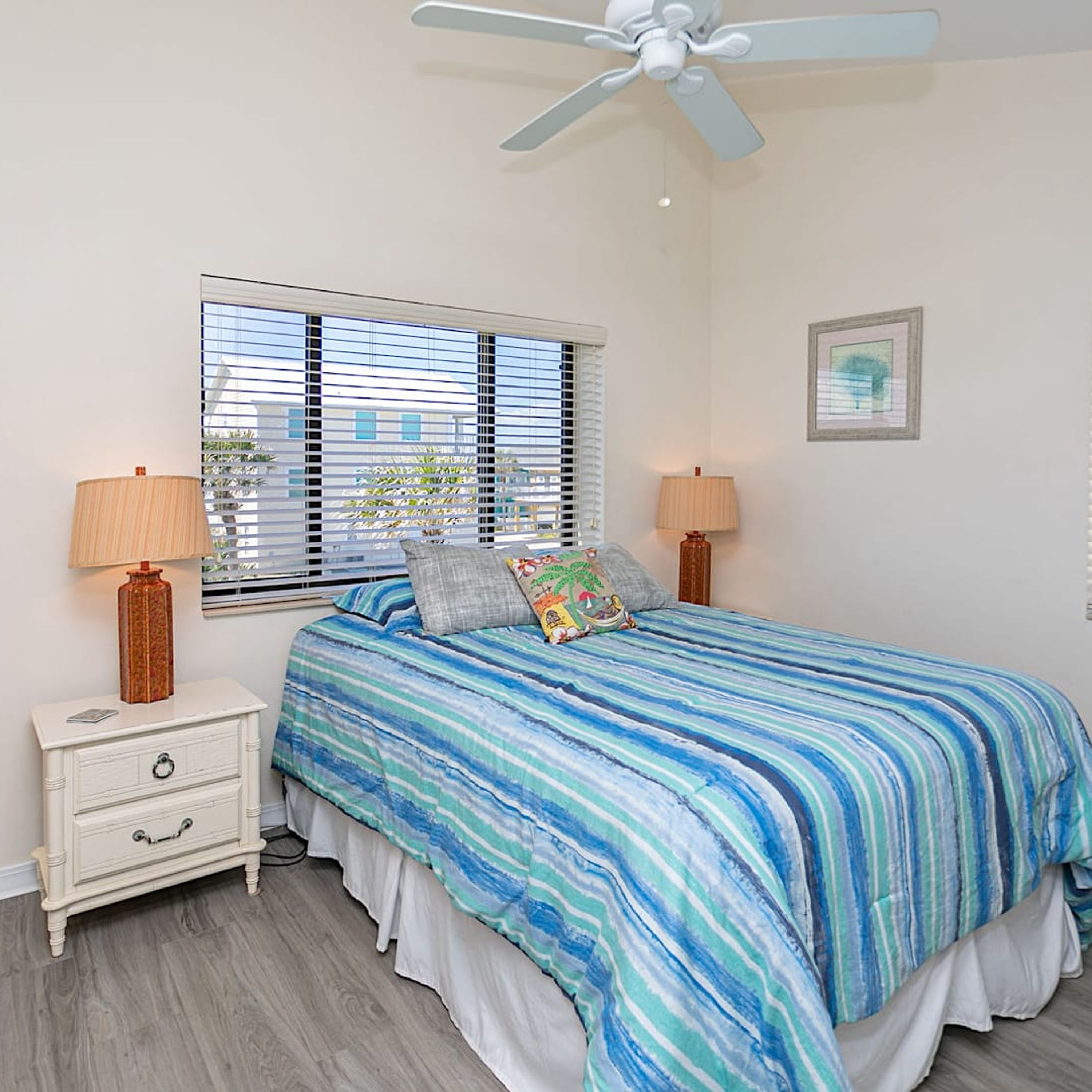 A bedroom with a bed, two bedside tables, a ceiling fan, a window with blinds, and a framed picture on the wall.