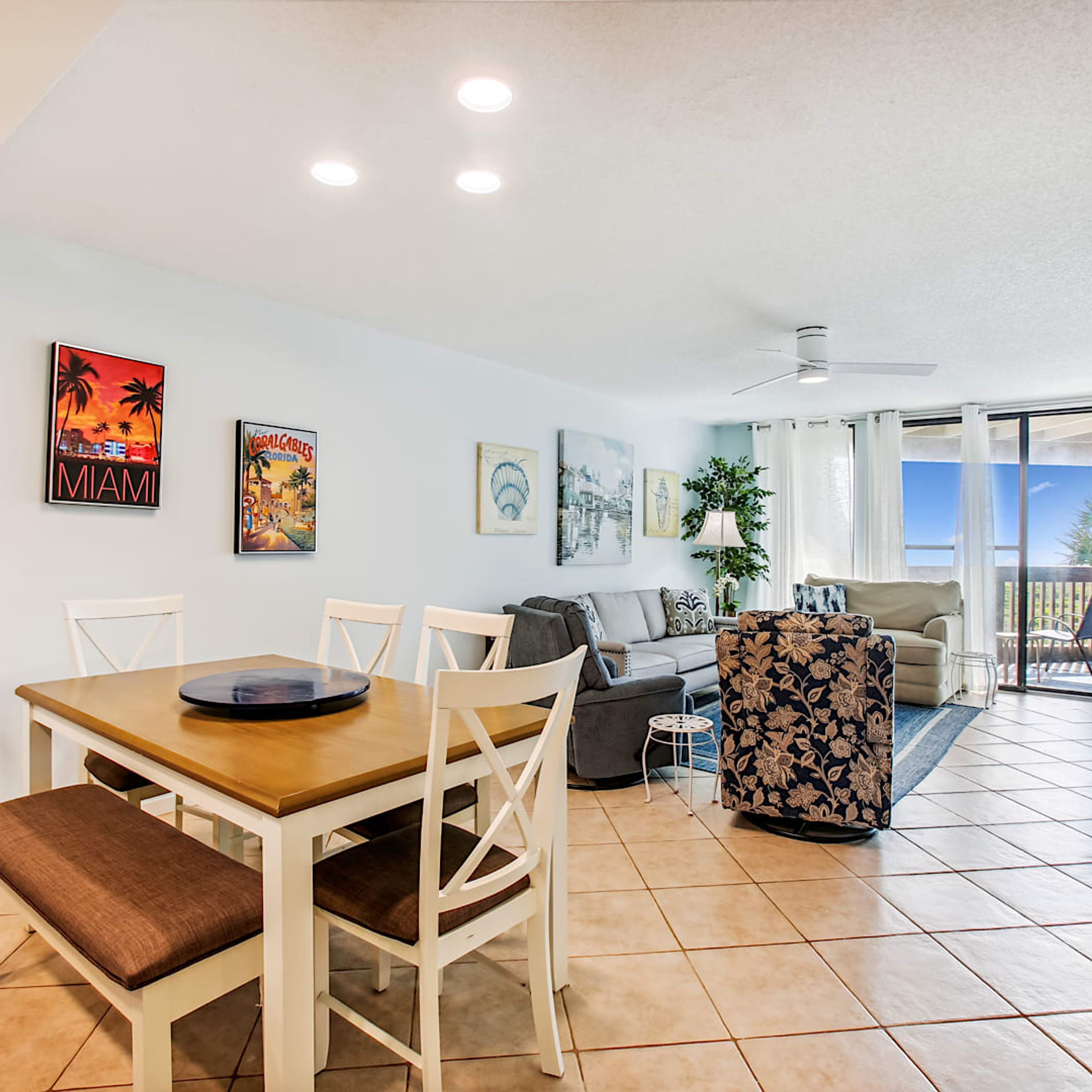 A spacious living area with a dining table, chairs, a sofa, and a patterned armchair. There are ceiling lights, a ceiling fan, and a view through a glass door to the outside.