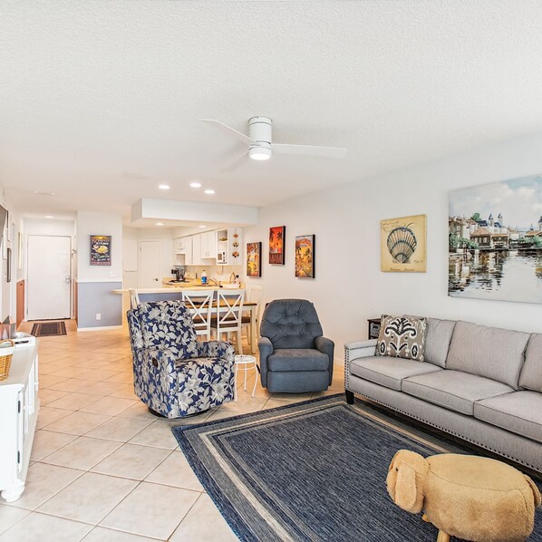A spacious living room with a sofa, a blue armchair, and a grey armchair. There is a dining area with a table and chairs in the background.