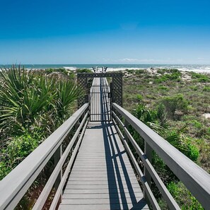 Une promenade en bois menant à une plage, avec une barrière à l’entrée, le tout entouré de verdure.