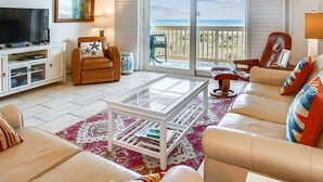 Ein Wohnzimmer mit Fernseher, Deckenventilator, Sofa, Couchtisch und Meerblick durch große Fenster.