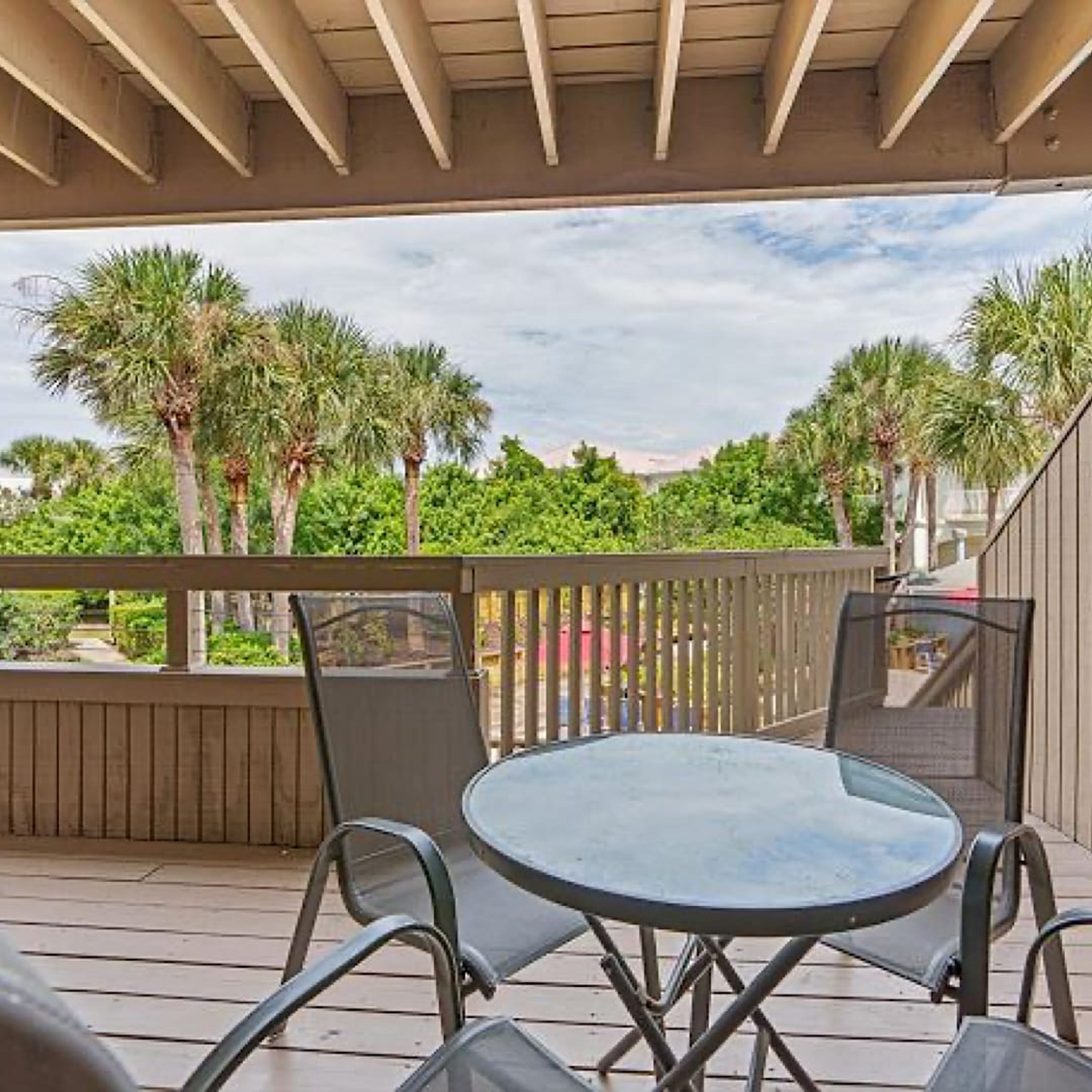 An outdoor patio with a round glass table and metal chairs, surrounded by a wooden deck and a metal railing.