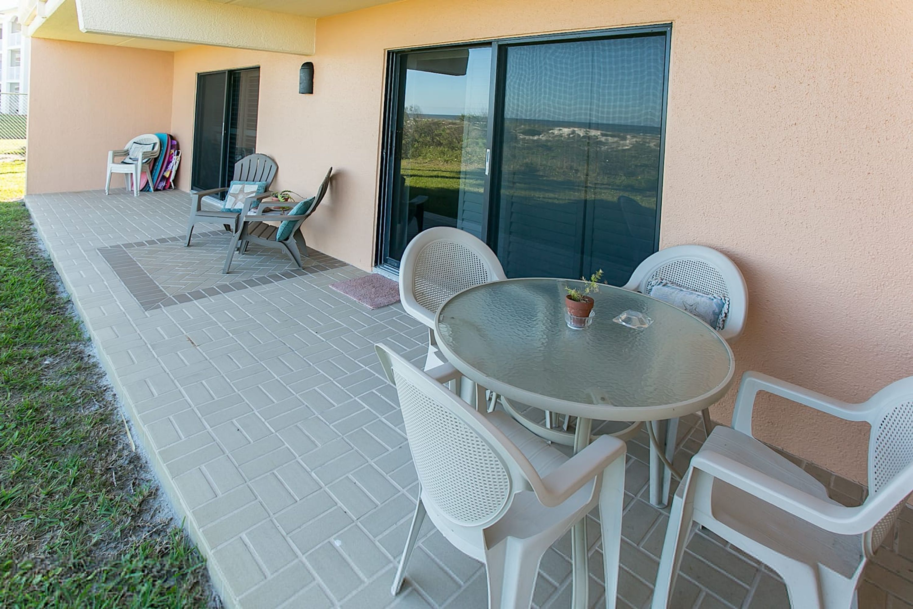Un patio con una mesa redonda y cuatro sillas, un ventanal corredero, y un pequeño jardín con una silla y una mochila.