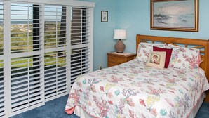 A bedroom with a bed, a nightstand, a lamp, a ceiling fan, and a window with blinds. - Sea Haven 221 Oceanfront Pool Comfort (St. Augustine)