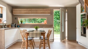 A modern kitchen with wooden cabinets, a dining table with chairs, and a view of greenery outside. - Sunnybrook in Shaldon (Teignmouth)