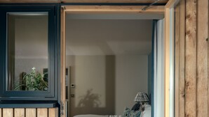 A bedroom with a wooden exterior, a bed with white bedding, a lamp, and a view of a plant outside. - Sunnybrook in Shaldon (Teignmouth)