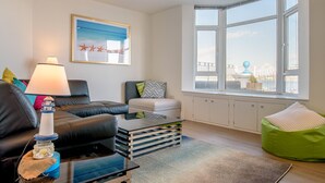 A modern living room with a black leather sofa, a glass coffee table, a green bean bag, and a large window with a view of the city. - Quayside View in Paignton (Paignton)