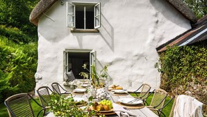 A dining table set outdoors with chairs, a white building in the background, and greenery around. - Weeke Brook in Chagford (Newton Abbot)