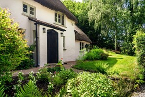 A white house with a black door and windows, surrounded by a lush garden with various plants and a thatched roof. - Weeke Brook in Chagford (Newton Abbot)