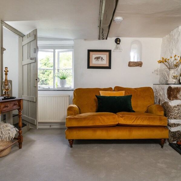 A room with a mustard-colored sofa, a small table with a lamp, a radiator, and a window with a view of greenery. - Weeke Brook in Chagford (Newton Abbot)