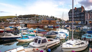 A harbor with various boats docked, a building with a traditional facade, and a hillside in the background. - Two Guns in Kingswear (Dartmouth)
