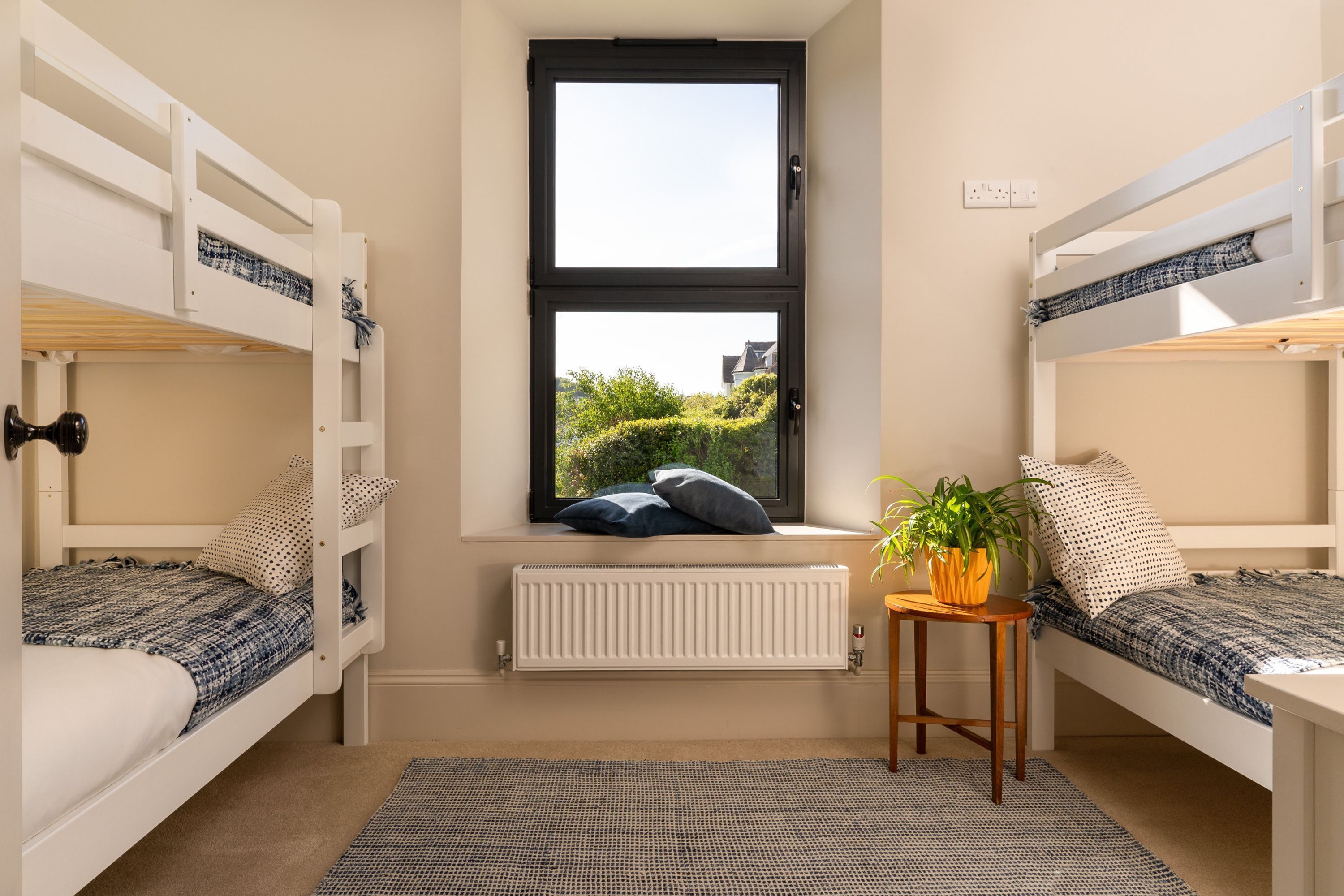 Ein Zimmer mit zwei Etagenbetten, einem Fenster mit Blick auf Grünflächen, einem Heizkörper und einem kleinen Tisch mit einer Pflanze.