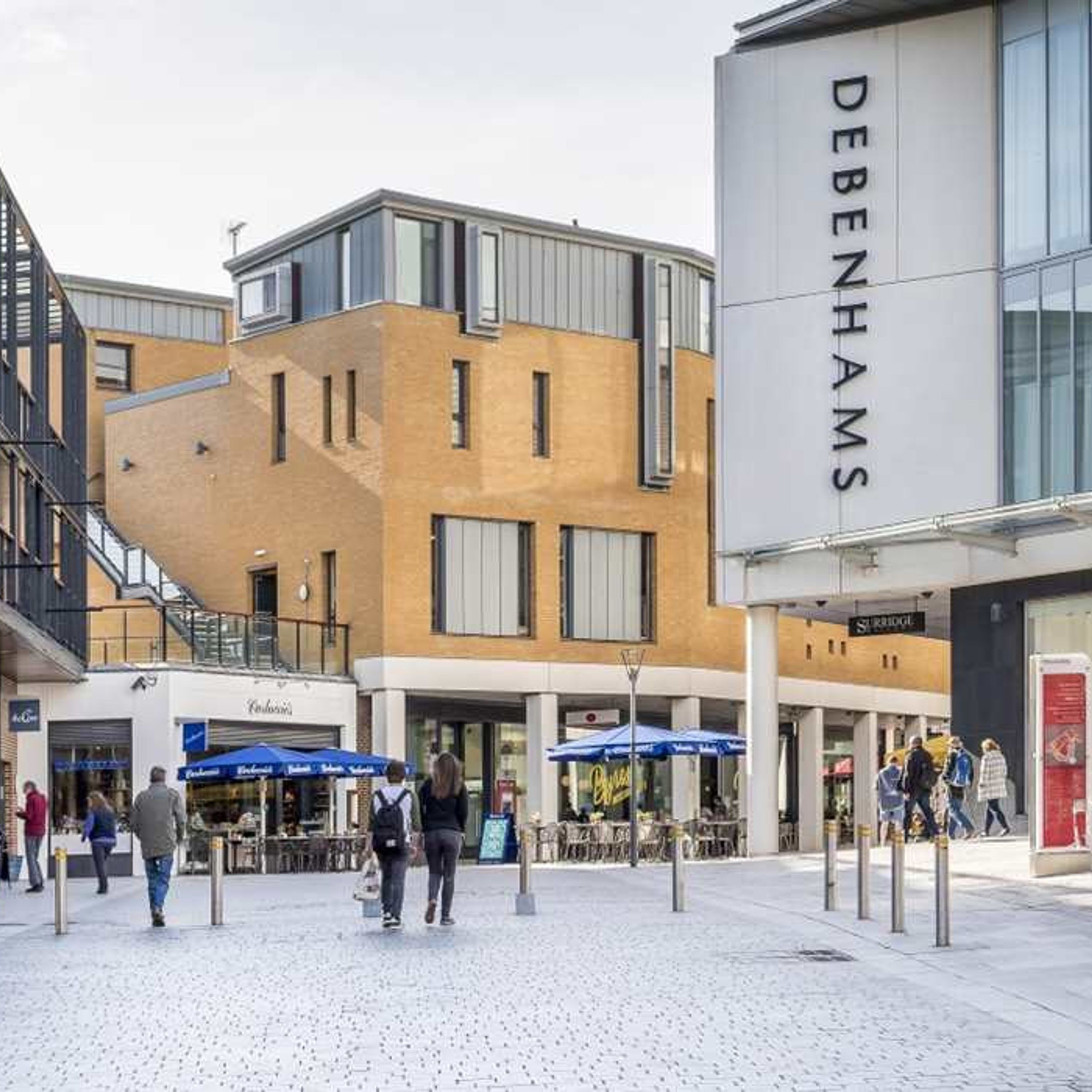 A modern urban square with a Debenhams store, outdoor seating, and a staircase leading to an upper level.