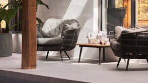 A modern outdoor seating area with wicker furniture and a table, surrounded by large windows and a potted plant. - Swallows End in Lower Ashton (Newton Abbot)