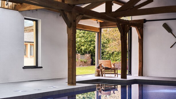 A modern indoor swimming pool with wooden beams, a clear glass wall, and a view of a garden. - Swallows End in Lower Ashton (Newton Abbot)