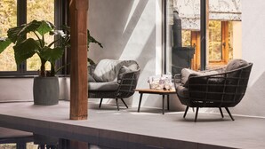 A modern outdoor seating area with wicker chairs and a small table, a potted plant, and a view of the outdoors through large windows. - Swallows End in Lower Ashton (Newton Abbot)