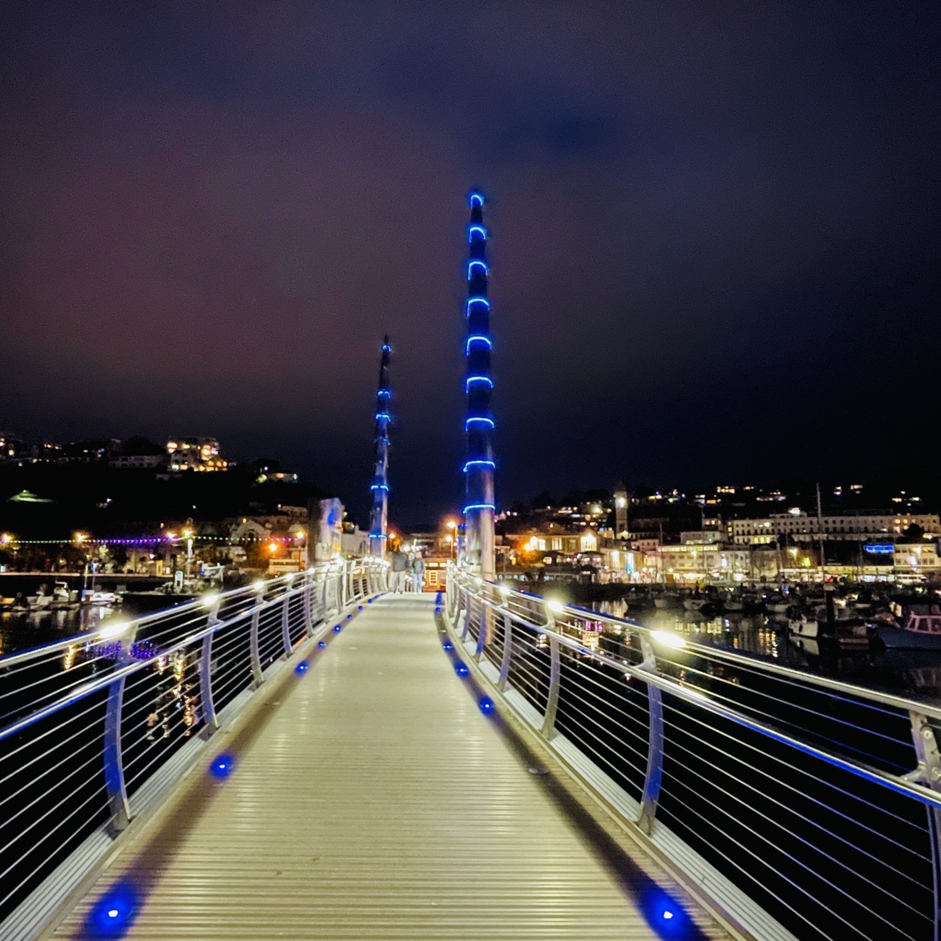 A bridge with railings and blue lights, leading to a cityscape with buildings and a body of water.