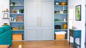 A room with a teal sofa, a wooden coffee table, a blue wardrobe with shelves, and a small table with a lamp.