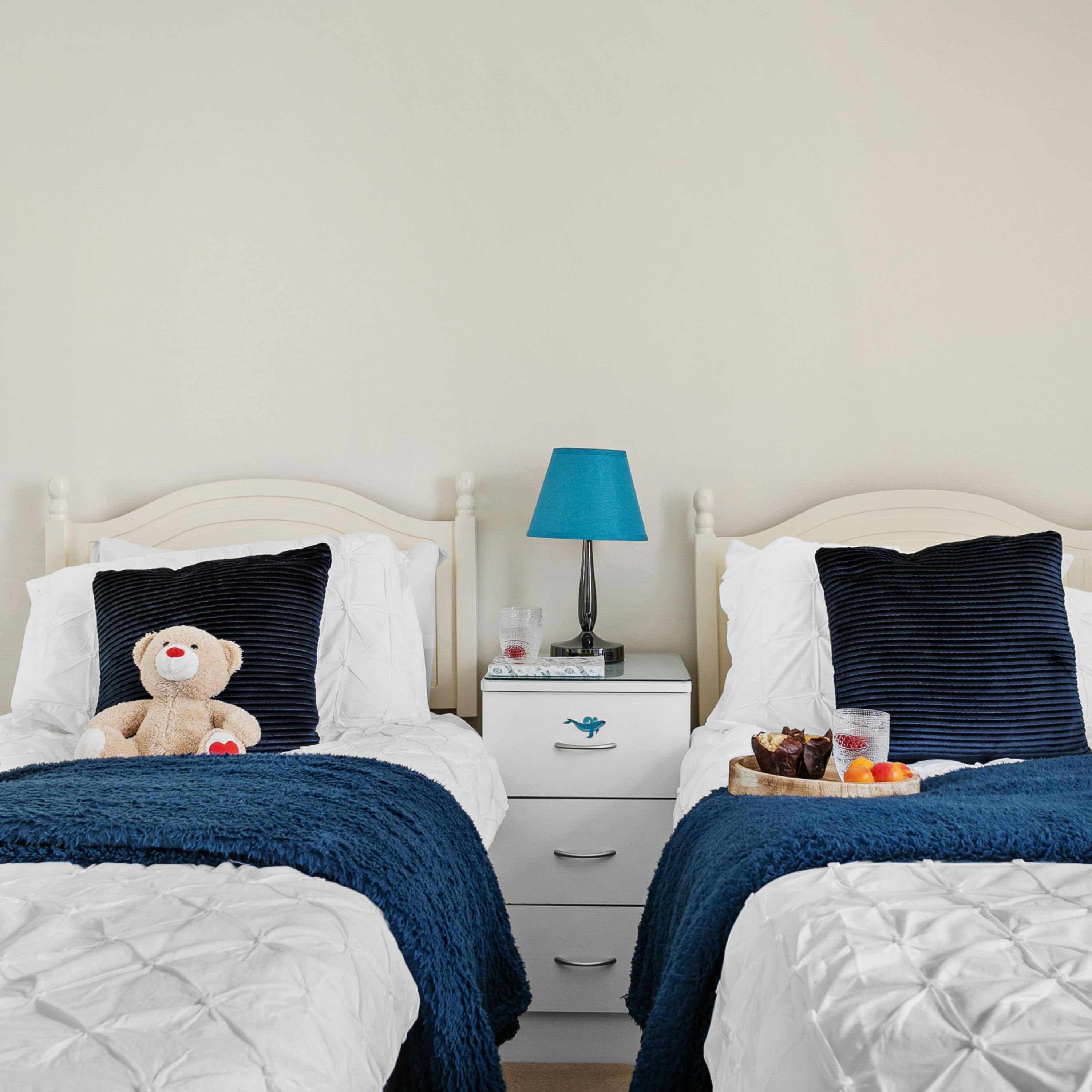 A twin bedroom with two beds, each with a white headboard and a dark blue blanket. There is a nightstand with a lamp between the beds, and a small table with a fruit bowl and a glass of water on the right side of the room.