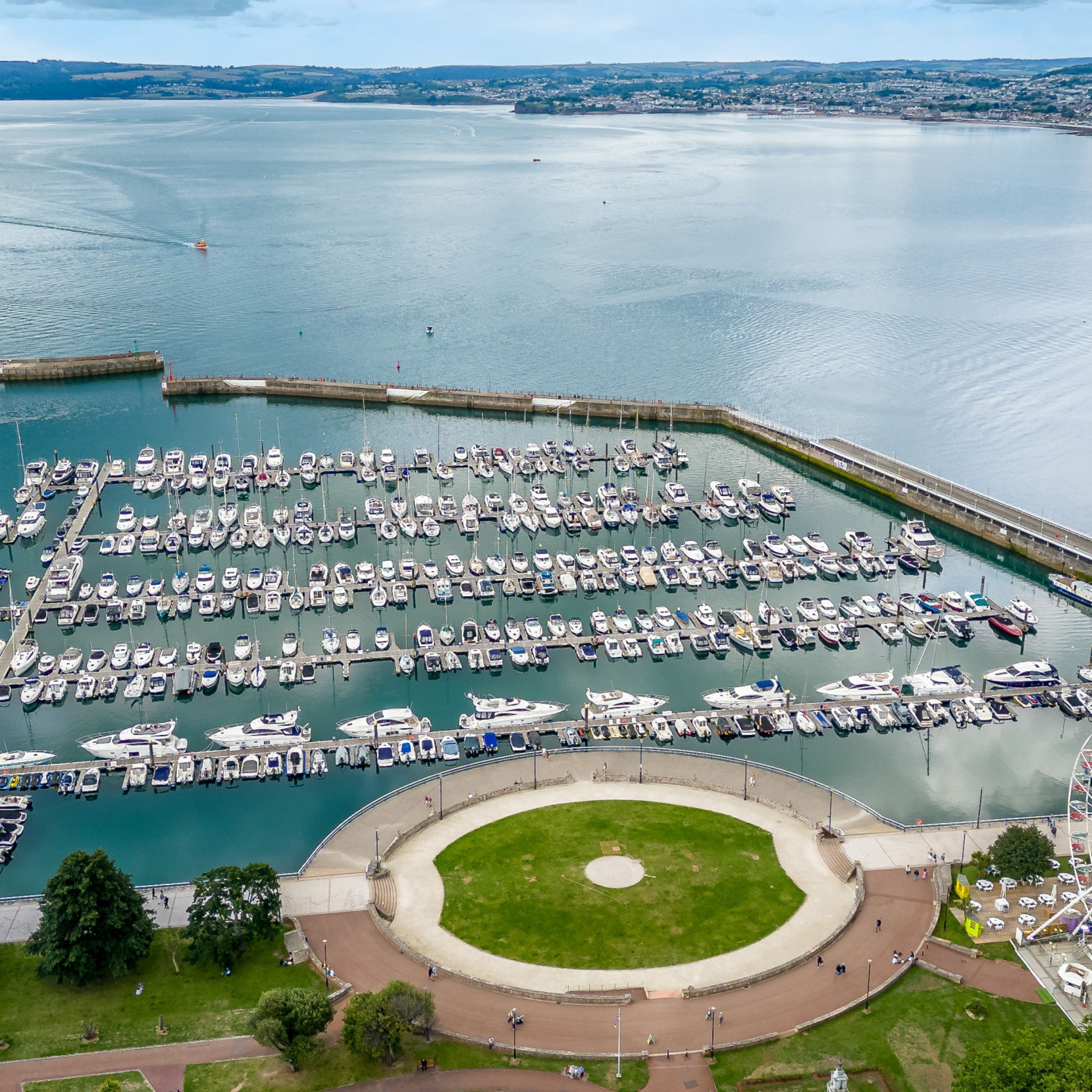An aerial view of a marina with numerous boats docked, a circular park area, and a distant cityscape.