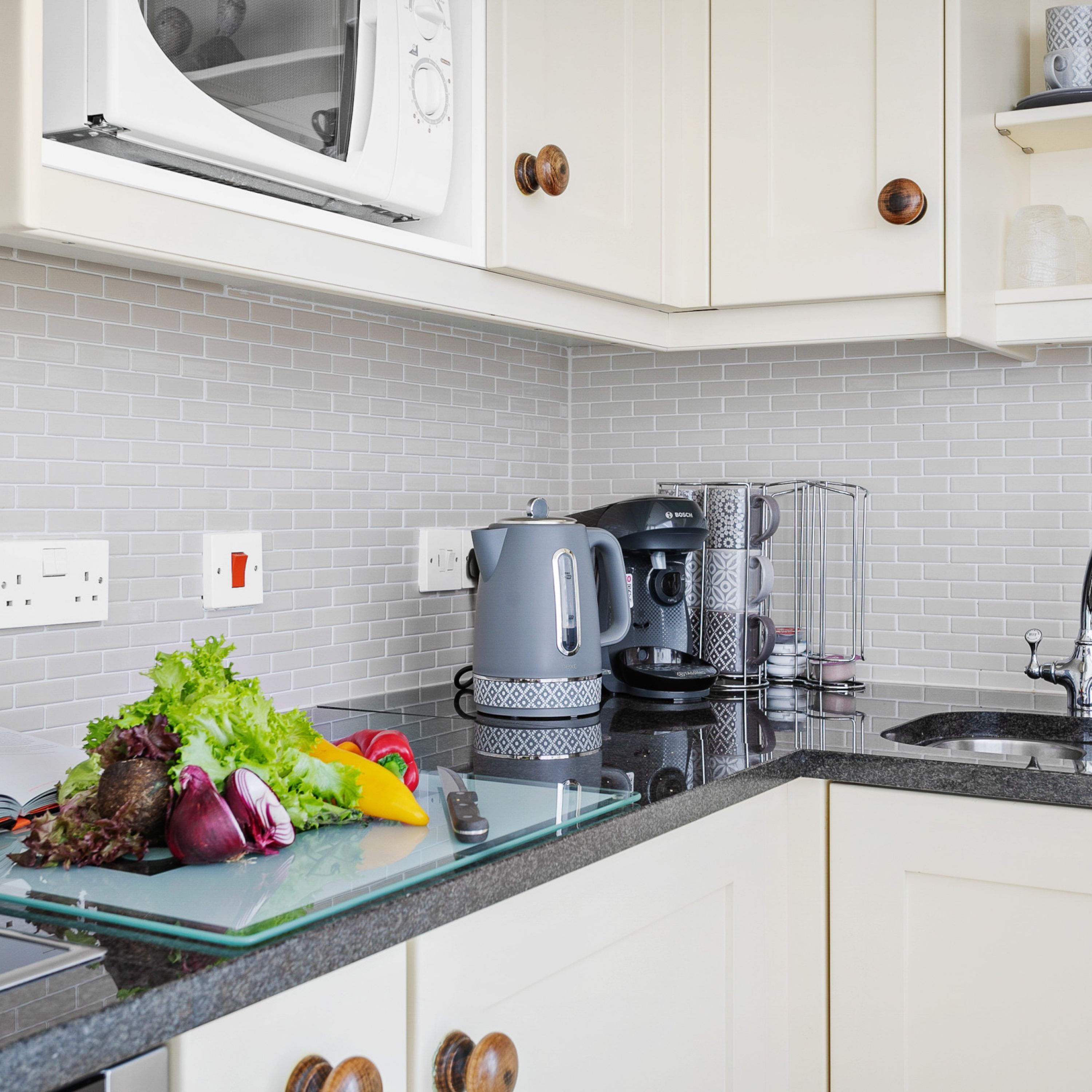 Cucina con mobili bianchi, un forno a microonde, una macchina da caffè e un tagliere di vetro con verdure.