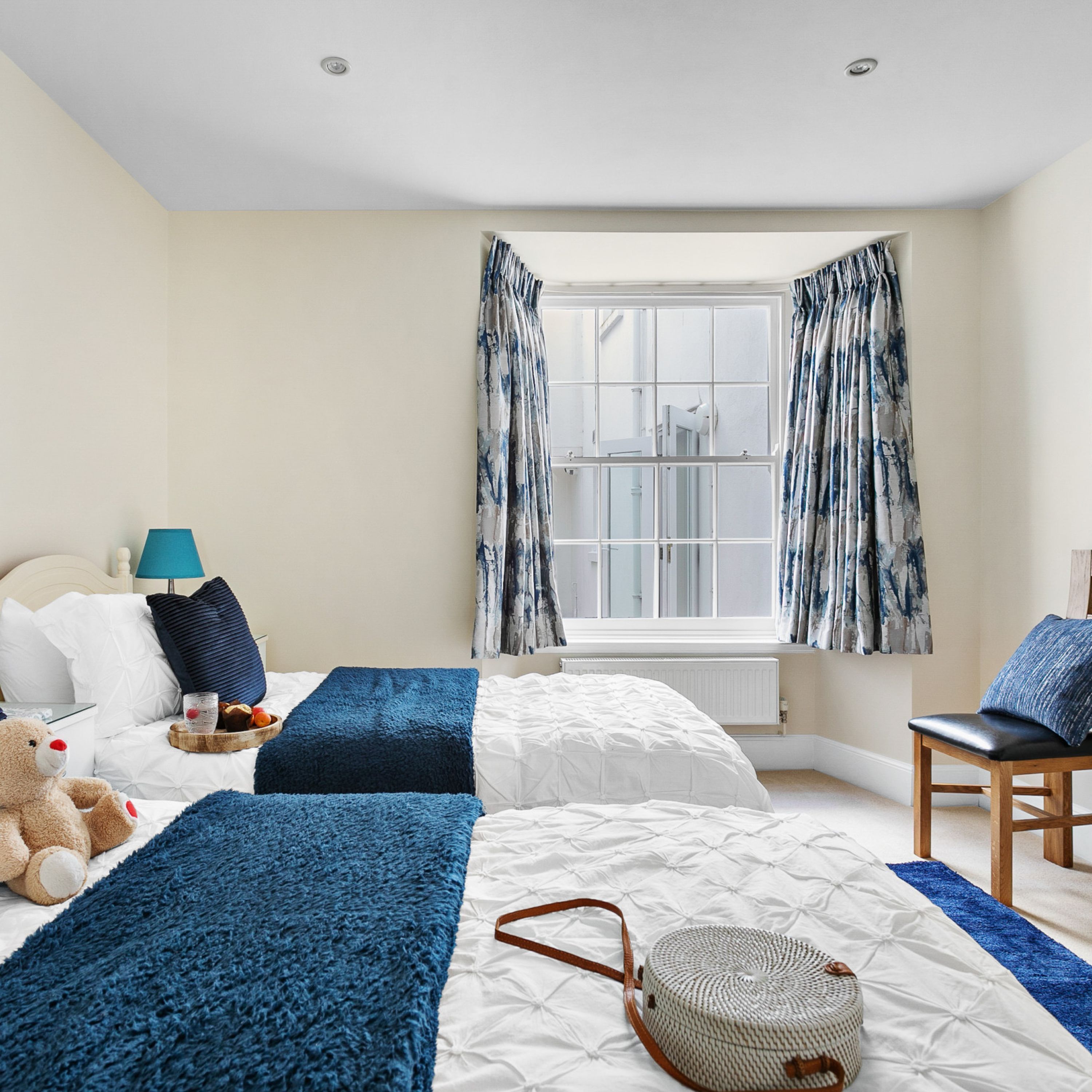 Une chambre d’hôtel avec deux lits, une fenêtre avec des rideaux, une lampe, un ours en peluche et un panier.