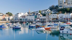 Un port de plaisance où sont amarrés différents types de bateaux, entouré de bâtiments aux styles architecturaux variés, dont une église dont le clocher est particulièrement imposant.