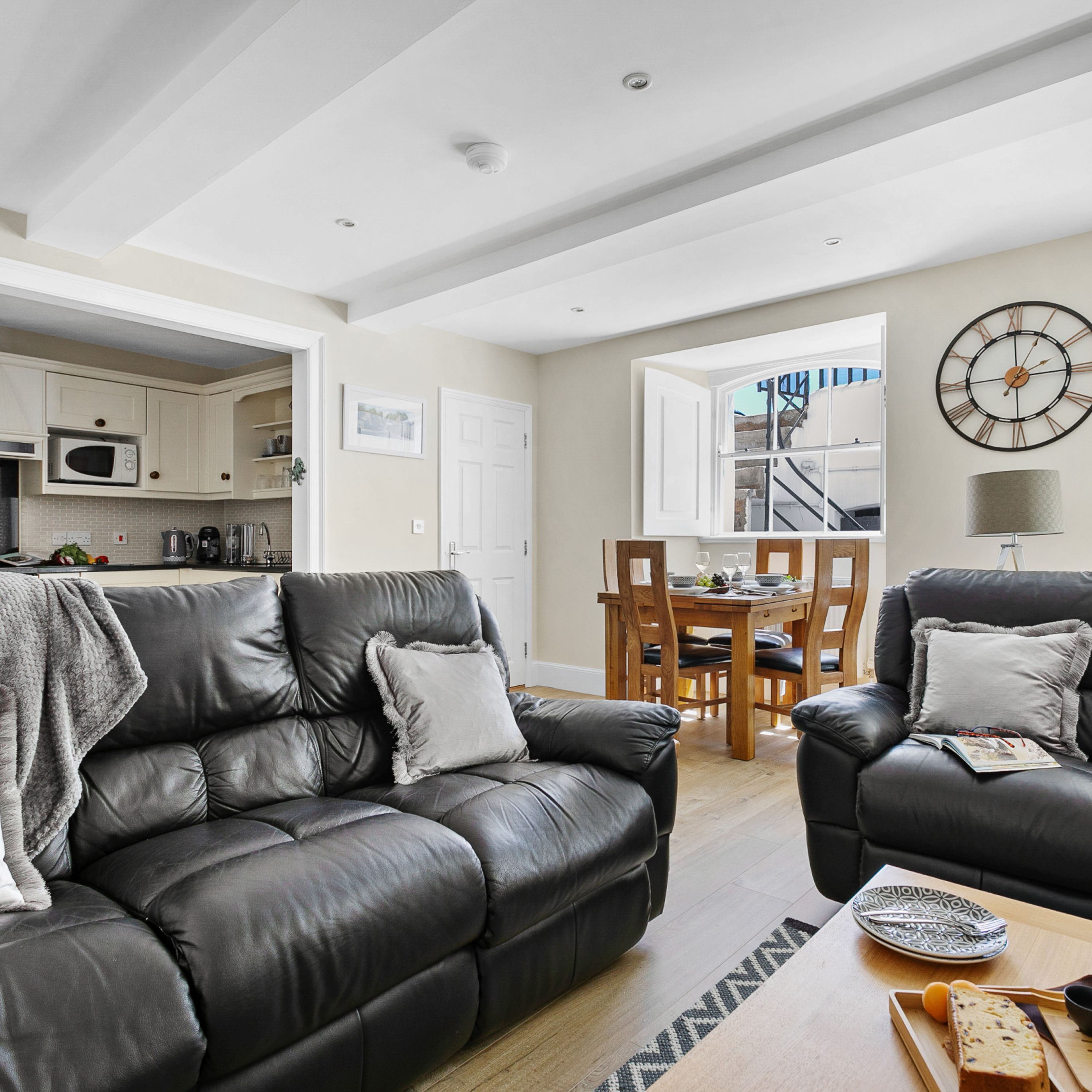A modern living room with a black leather sofa, a dining table with chairs, and a kitchen in the background.