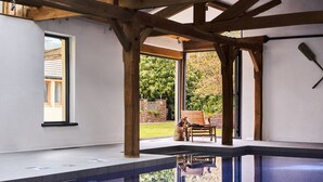 A modern indoor pool with a clear view of the outdoors, wooden beams, and a patio area with a chair. - Swifts Return in Lower Ashton (Newton Abbot)