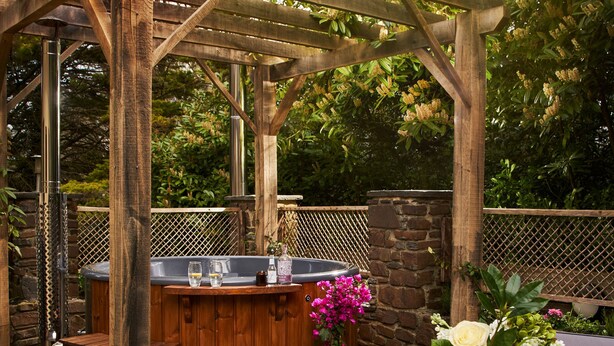 An outdoor patio with a wooden pergola, a hot tub, and a dining table set with a plate of food and a glass of wine. - Swifts Return in Lower Ashton (Newton Abbot)