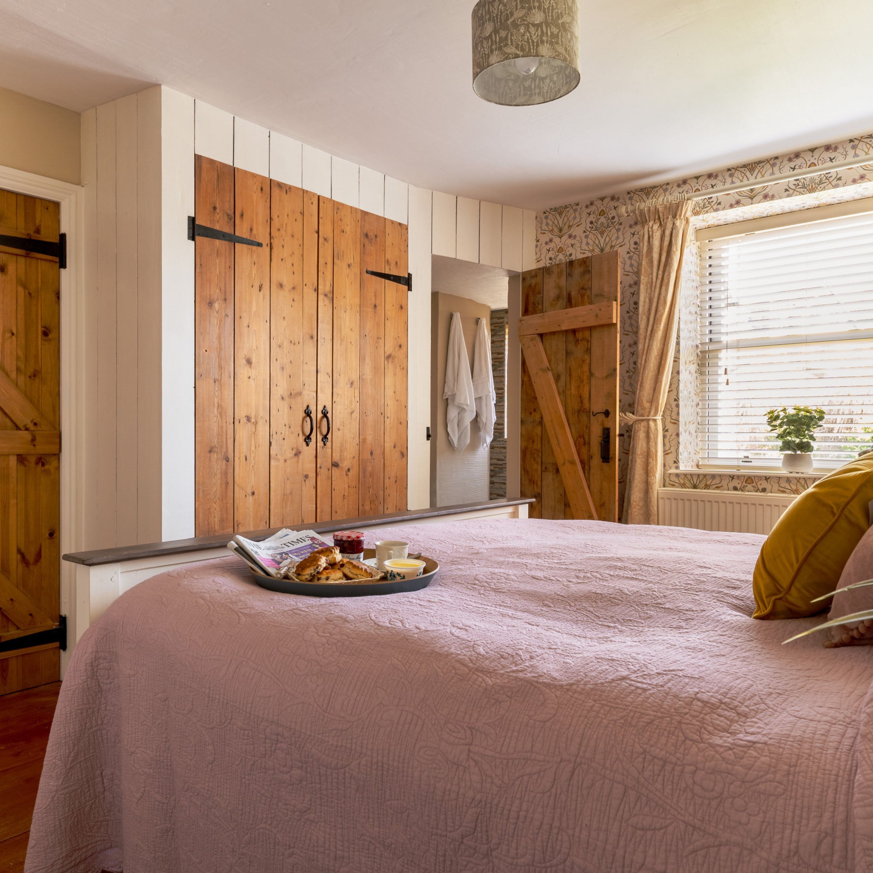 Un dormitorio con cama, armario de madera, ventana con cortinas y una bandeja con comida sobre la cama.