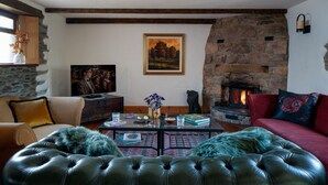 A cozy living room with a stone fireplace, a comfortable sofa, a leather ottoman, and a coffee table with books and decorative items. - Old Coach House in Berry Pomeroy (Totnes)