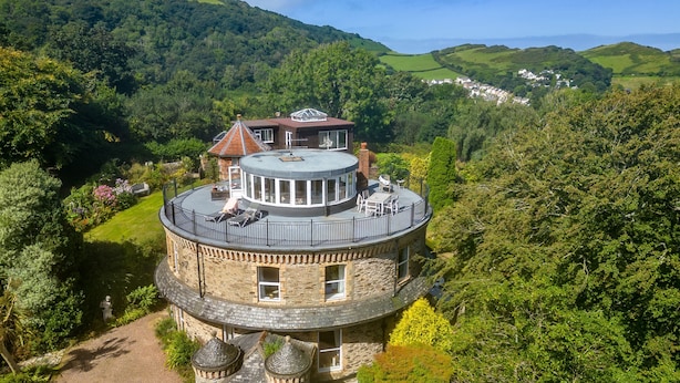 A stone tower house with a circular balcony, surrounded by lush greenery and hills. - The Round House in Ilfracombe (Ilfracombe)
