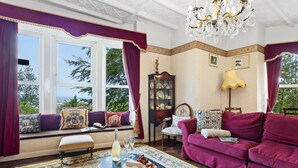 A room with a window seat, a sofa, a table with a bottle and glasses, and a chandelier. - The Round House in Ilfracombe (Ilfracombe)