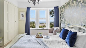 A bedroom with a bed, a chair, a window with a view, a chandelier, and a wardrobe. - The Round House in Ilfracombe (Ilfracombe)