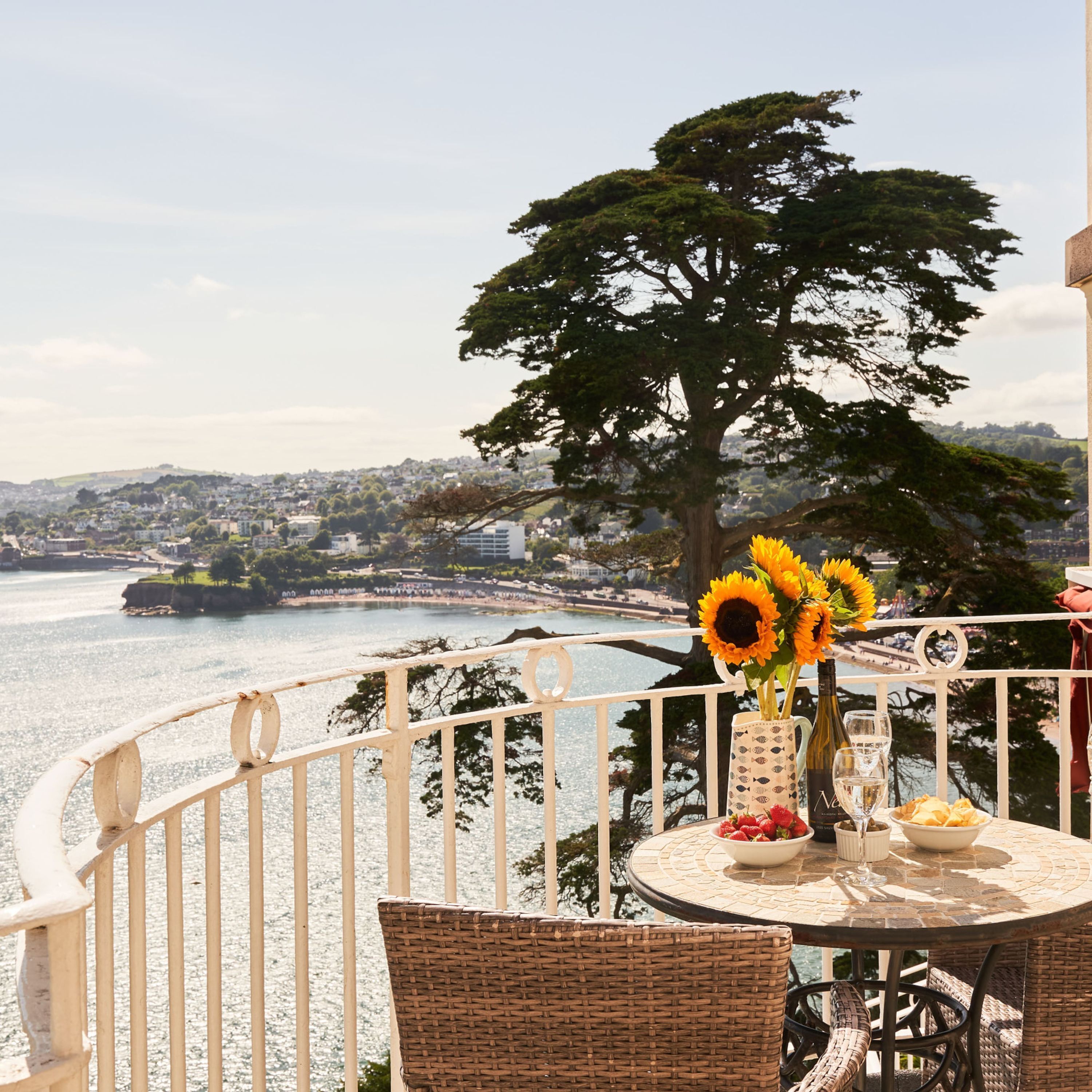 A balcony with a table set for dining, overlooking a body of water and a cityscape.