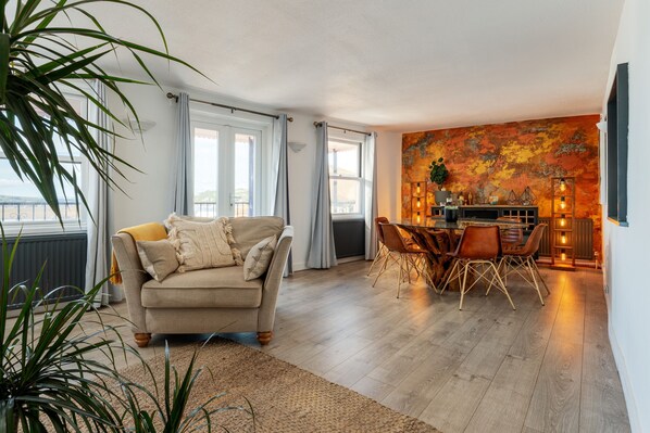 Una sala de estar con un sofá color beige, un comedor con una mesa y sillas de madera, y un ventanal con cortinas.